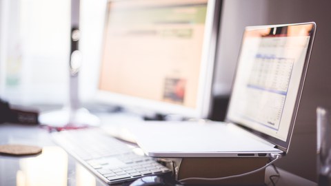 Arbeitsplatz mit einem Pc und einem Laptop rechts daneben, der auf einem dicken Buch steht.