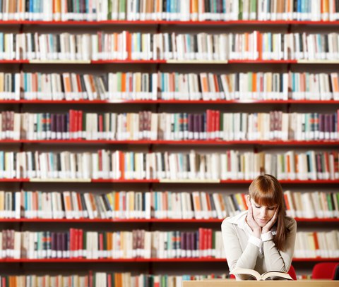 Foto einer jungen Studentin, die vor einer Bücherwand an einem Tisch sitzt und ein Buch liest.