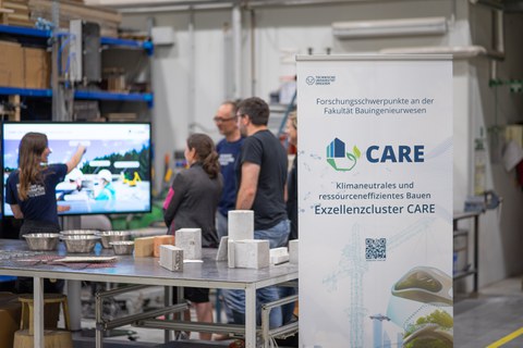 Interessierte Menschen hören sich eine Präsentation einer jungen Wissenschaftlerin vor einem großen Bildschirm an. Auf dem Tisch im Vordergrund liegen ein paar Exponate aus Beton und alternative Bewehrungen