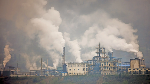 Industrial plant with several buildings and chimneys from which a lot of smoke or vapour rises.