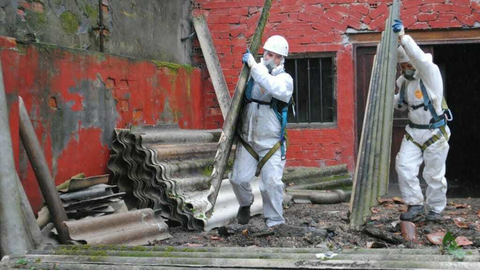 Two men in white full-body protective suits carrying asbestos roof panels