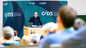 Prof. Dr. Lars Bernard am Rednerpult vor den CIDS-Beschäftigten (Fotoaufnahme in einem steilen Hörsaal, mit einer Vielzahl von Rückenansichten der Zuhörenden im Vordergrund und Porf. Bernard im Hintergrund vor einer Wand im Corporate Design des CIDS).)