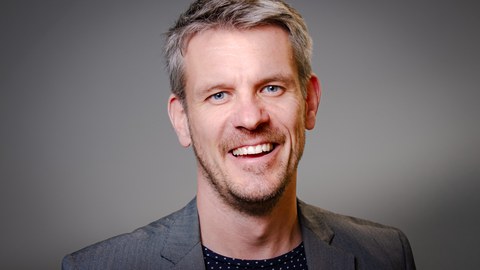 weißer Mann mittleren Alters, graues Jacket, lackt in die Kamera
