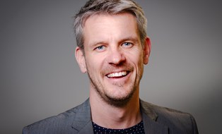 weißer Mann mittleren Alters, graues Jacket, lackt in die Kamera