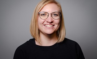 Blonde woman with black glasses, black shirt, smiling at the camera