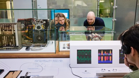 Zwei Personen betrachten einen Ausstellungsbereich. Eine Person im Vordergrund rechts hört einen Audiobeitrag der Ausstellung an.