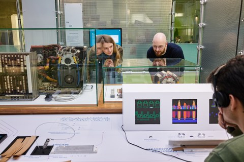 Zwei Personen betrachten einen Ausstellungsbereich. Eine Person im Vordergrund rechts hört einen Audiobeitrag der Ausstellung an.