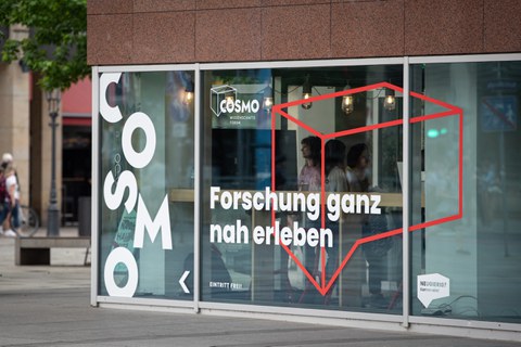 Außenaufnahme des COSMO Wissenschaftsforums im Kulturpalast. Auf den Scheiben das COSMO-Logo und der Schriftzug "Forschung ganz nah erleben".