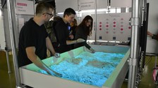 Drei Besuchende der Ausstellung entdecken die Funktionsweise von Schwarmverhalten am Exponat der interaktiven Sandbox.