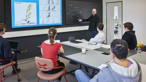 In einem modernen Seminarraum hält ein Dozent eine wissenschaftliche Präsentation vor einer Gruppe von fünf Teilnehmenden. Er steht vor einer Tafel, auf der eine mathematische Gleichung skizziert ist, und zeigt auf eine Formel. Neben ihm ist ein großer Bildschirm mit einer grafischen Darstellung genomischer und geografischer Daten zu sehen. Die Zuhörerinnen und Zuhörer, darunter eine Frau in einem roten Oberteil und vier Männer in legerer Kleidung, sitzen an einem Tisch mit Laptops, Notizbüchern und Kaffeetassen. Der Raum ist hell und funktional eingerichtet, mit einer schwarzen Wand als Kontrast.