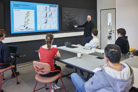 In einem modernen Seminarraum hält ein Dozent eine wissenschaftliche Präsentation vor einer Gruppe von fünf Teilnehmenden. Er steht vor einer Tafel, auf der eine mathematische Gleichung skizziert ist, und zeigt auf eine Formel. Neben ihm ist ein großer Bildschirm mit einer grafischen Darstellung genomischer und geografischer Daten zu sehen. Die Zuhörerinnen und Zuhörer, darunter eine Frau in einem roten Oberteil und vier Männer in legerer Kleidung, sitzen an einem Tisch mit Laptops, Notizbüchern und Kaffeetassen. Der Raum ist hell und funktional eingerichtet, mit einer schwarzen Wand als Kontrast.