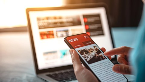 Foto: Eine Person hält ein Smartphone mit einer geöffneten Nachrichten-App vor einem Laptop mit Nachrichtenwebseite