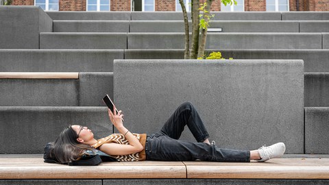 Bild einer Studierenden, die auf einer Treppe liegt und an einem Tablet scrollt.