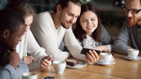 Fünf junge Erwachsene sitzen mit Kaffee und Kuchen am Tisch und schauen auf ihre Handys
