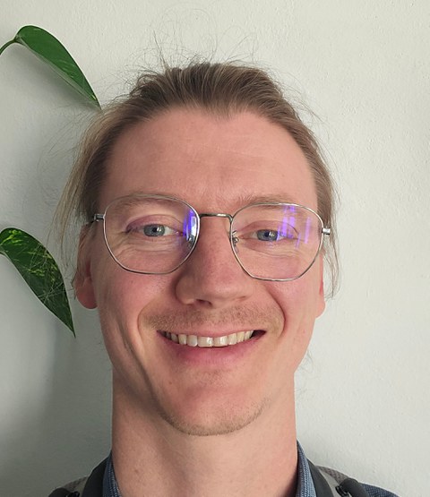 Das bild zeigt Dr. Pascal Klamser in einer frontalen Ansicht. Er hat blonde lange Haase zu einem Zopf gebunden und trägt eine runde Brille.