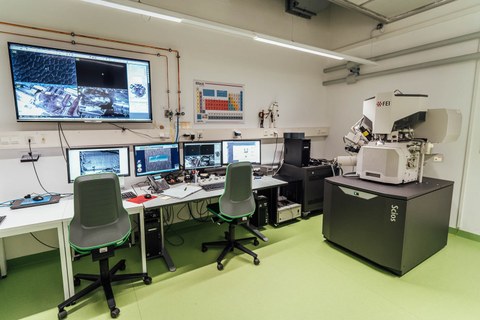 Room with a machine standing in the right corner and desks along the adjacent wall. 4 screens are standing on the desks with two chairs in front of them. On the wall is a larger screen.