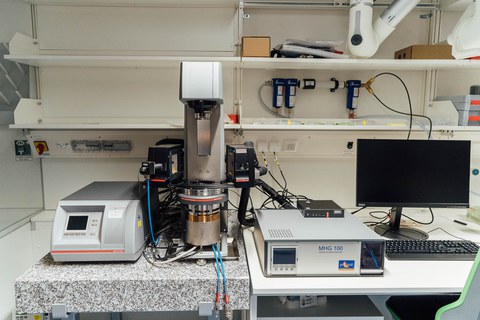 A desk in front of a wall with empty shelf boards. On the desk a computer is visible on the right side of a machine and 2 electrical devices conecte to it via multiple cables.