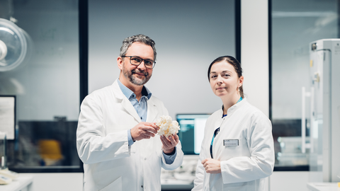 Ein Foto. Zwei Personen in weißen Laborkitteln posieren für ein Foto in einer laborähnlichen Umgebung. Die Person links hält eine weiße, körnige Struktur, etwa so groß wie ein Handball, in den Händen.