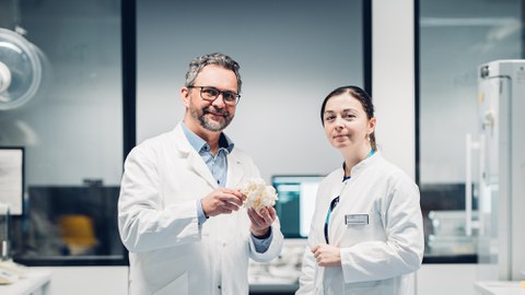 A photo. Two people wearing white labcoats pose for a picture in a laboratory-like setting. The person on the left holds a white grainy structure, the size of a handball ball, in his hands.