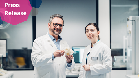 Ein Foto. Zwei Personen in weißen Laborkitteln posieren für ein Foto in einer laborähnlichen Umgebung. Die Person links hält eine weiße, körnige Struktur, etwa so groß wie ein Handball, in den Händen.