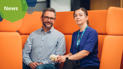 Ein Foto. Zwei Personen sitzen nebeneinander auf einem futuristischen orangefarbenen Sofa. Die Person auf der rechten Seite hält einen weißen Gegenstand mit unregelmäßiger Form in der Hand.