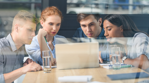 The photo shows four young people behind a glass front, sitting together at a table and concentratedly looking into a laptop.