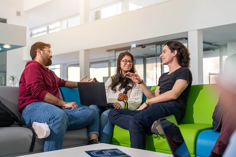 Ein Foto. Drei Personen sitzen auf einem Ecksofa und unterhalten sich miteinander. Die Person in der Mitte hat einen Laptop auf ihrem Schoß liegen. Die Person links sowie die Person in der Mitte schauen zur Person rechts. Alle haben fröhliche Gesichter und sind in ein lebhaftes Gespräch vertieft.