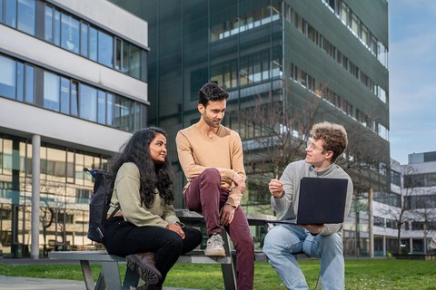 A photo. Three people are sitting on a bench outside, talking to each other. There is grass and glass buildings in the background. The person on the right has a laptop in his hand, presenting content to the other two people. The person on the left is looking towards the person on the right. The person in the middle is looking towards the laptop. Everybody has focused faces and are engaged in a lively conversation.