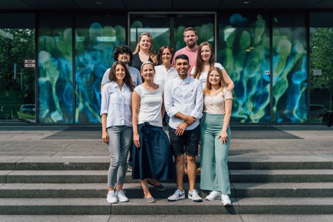 Group picture of 9 people standing in three rows on stairs in front of glass doors with a blue and green print on them.