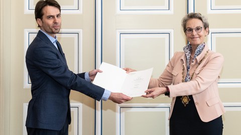 Zwei Personen. Ein Mann auf der linken Seite, der einen Anzug trägt. Eine Frau auf der rechten Seite, die ein formelles Outfit trägt. Gemeinsam halten sie eine Mappe in der Hand.