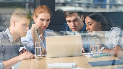 Das Foto zeigt vier junge Personen hinter einer Glasfront, die gemeinsam an einem Tisch sitzen und konzentriert in einen Laptop schauen.