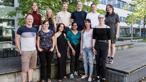 The 12 members of the Zeißig research group standing in 2 rows of 6 people each, with a tree and a white building with big windows in the background.