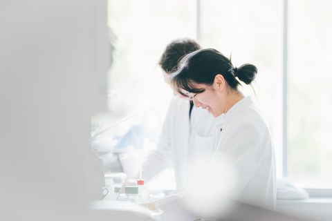A photo. Two people in lab coats. They are standing at a table, and in front of them, a cabinet is hanging. This table and cabinet are in the left half of the picture and are out of focus. Various laboratory utensils are on the table. The two people are working with these utensils.