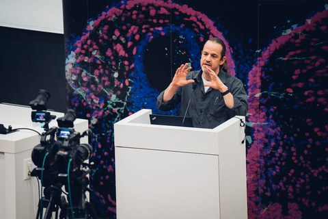 A man stands in front of a colorful background. He speaks to the microphones and cameras in front of him. 