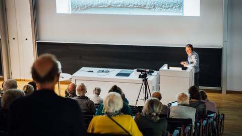 Prof. Dr. Marius Ader steht an einem Pult und redet vor einem Publikum in einem Hörsaal. Hinter ihm wird eine Präsentation per Beamer an die Wand projeziert. In der Mitte des Raumes steht eine Kamera zur Live-Übertragung des Vortrages.