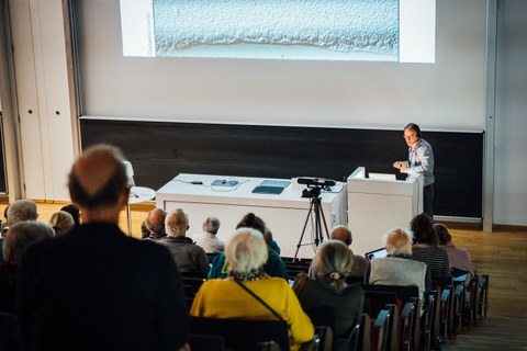 Prof. Dr. Marius Ader stands at a podium and speaks to an audience in a lecture hall. Behind him, a presentation is projected onto the wall via beamer. In the middle of the room stands a camera for the live transmission of the lecture.