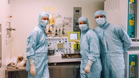 Three people wearing protective suits, gloves, masks, and hats pose in front of a machine in a laboratory setting. The machine has IV-bags connected to it.