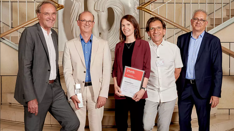 A group photo with five people. The two people in the middle hold a diplom and an award that looks like a block of metal and glass.