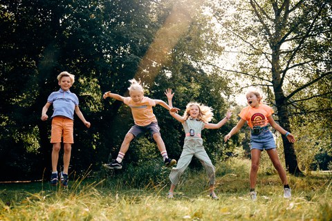 Ein Foto. Vier Kinder springen gleichzeitig vor einer Kulisse aus dichten grünen Bäumen und einer sonnenbeschienenen Wiese. Von links nach rechts tragen sie ein blau gestreiftes Hemd mit orangefarbenen Shorts, ein hellorangefarbenes Shirt, einen gemusterten blaugrünen Jumpsuit und ein leuchtend orangefarbenes Shirt mit Jeans-Shorts. Jedes Kind ist in einer individuellen, bewegungsreichen Sprungpose in der Luft eingefroren. Ein heller Sonnenstrahl verläuft diagonal von oben rechts durch die Bildmitte.