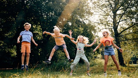 Ein Foto. Vier Kinder springen gleichzeitig vor einer Kulisse aus dichten grünen Bäumen und einer sonnenbeschienenen Wiese. Von links nach rechts tragen sie ein blau gestreiftes Hemd mit orangefarbenen Shorts, ein hellorangefarbenes Shirt, einen gemusterten blaugrünen Jumpsuit und ein leuchtend orangefarbenes Shirt mit Jeans-Shorts. Jedes Kind ist in einer individuellen, bewegungsreichen Sprungpose in der Luft eingefroren. Ein heller Sonnenstrahl verläuft diagonal von oben rechts durch die Bildmitte.