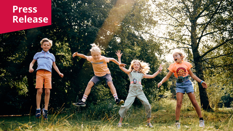 Ein Foto. Vier Kinder sind mitten im Sprung vor einer Kulisse aus dichten grünen Bäumen und einer sonnenbeschienenen Wiese abgebildet. Sie tragen farbenfrohe Sommerkleidung, darunter gestreifte und orangefarbene Shirts, Shorts sowie einen gemusterten Jumpsuit. Jedes Kind befindet sich in einer dynamischen Sprungpose mit ausgestreckten Armen und Beinen. Ein heller Sonnenstrahl verläuft diagonal von oben durch die Bildmitte. In der oberen linken Ecke befindet sich ein magentafarbenes rechteckiges Banner mit dem weißen Schriftzug „Press Release“