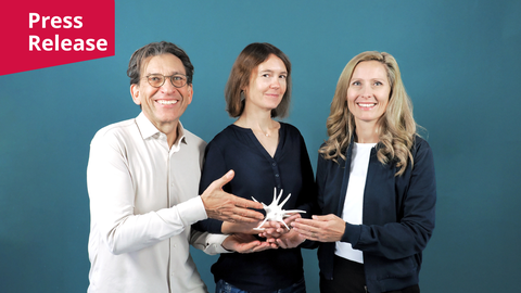 A photo. Portrait of three people smiling. They hold a white shape, looking like made of porcelain, together in their hands. In the upper left corner, there is a red shape with white text "Press Release."