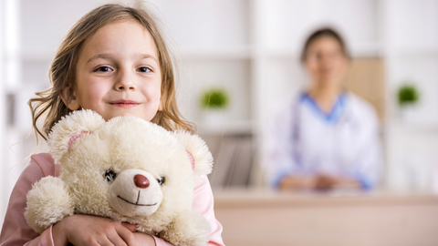 A photo. In the foreground, a young child with long light-brown hair and a slight smile is holding a cream-colored teddy bear tightly to their chest. The child is wearing a light pink long-sleeved shirt, and the teddy bear has a simple stitched smile and a brown nose. The background is blurred, showing a person in a white coat with a stethoscope, suggesting a clinical or office setting with white shelves and small green plants. The lighting is bright and even, highlighting the child and the bear as the central focus.