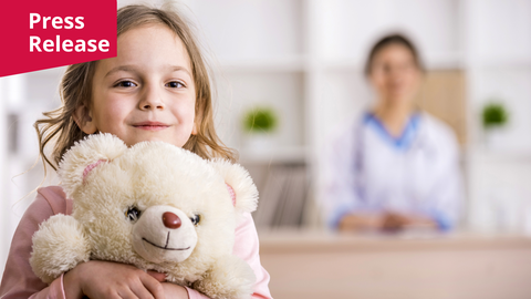 Ein Foto. Im Vordergrund hält ein junges Kind mit langen, hellbraunen Haaren und einem leichten Lächeln einen cremefarbenen Teddybären fest an seine Brust. Das Kind trägt ein hellrosa langärmeliges Oberteil, und der Teddybär hat ein einfach gesticktes Lächeln und eine braune Nase. Der Hintergrund ist unscharf und zeigt eine Person in einem weißen Kittel mit einem Stethoskop, was auf eine klinische Umgebung oder ein Büro mit weißen Regalen und kleinen grünen Pflanzen hindeutet. Die Beleuchtung ist hell und gleichmäßig, wodurch das Kind und der Bär als zentraler Fokus hervorgehoben werden. Ein rotes rechteckiges Banner mit dem weißen Wort „Press Release“ ist in der oberen linken Ecke platziert.