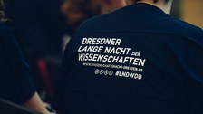 A photo of the back of a person. They are wearing a T-shirt with a print: „DRESDNER LANGE NACHT der WISSENSCHAFTEN“ under that, the internet address: „www.wissenschaftsnacht-dresden.de“. Below that, four logos of Instagram, facebook, X (former Twitter) und LinkenIn, besides that there’s the Hashtag: #LNDWDD.