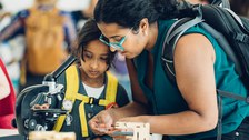 A photo of a person with their child at an old microscope, holding a glass container.