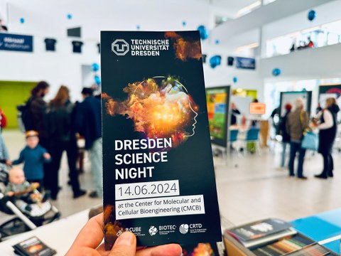 Eine Hand hält einen Flyer für die 'Dresdner Lange Nacht der Wissenschaften' an der Technischen Universität Dresden. Der Flyer zeigt ein abstraktes Bild eines menschlichen Profils mit einem lebendigen, bunten wolkenartigen Design im Kopf. Die Veranstaltungsdetails sind angegeben: '14.06.2024 im Center for Molecular and Cellular Bioengineering (CMCB)'. Im Hintergrund sieht man einen modernen, belebten Innenraum mit Menschen, die interagieren, und verschiedenen Ausstellungen in der Ferne.