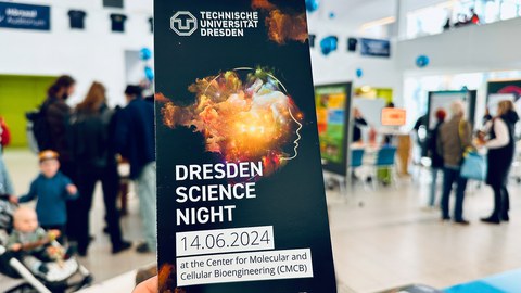 Eine Hand hält einen Flyer für die 'Dresdner Lange Nacht der Wissenschaften' an der Technischen Universität Dresden. Der Flyer zeigt ein abstraktes Bild eines menschlichen Profils mit einem lebendigen, bunten wolkenartigen Design im Kopf. Die Veranstaltungsdetails sind angegeben: '14.06.2024 im Center for Molecular and Cellular Bioengineering (CMCB)'. Im Hintergrund sieht man einen modernen, belebten Innenraum mit Menschen, die interagieren, und verschiedenen Ausstellungen in der Ferne.