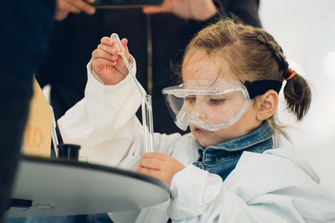 Ein Foto eines kleinen Kindes mit Laborkittel und Schutzbrille welches eine Pipette in ein Reagenzglas hält.