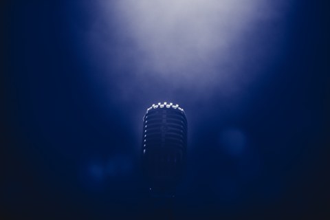 ein künstlerisches Bild eines Bühnenmikrofons. Das Mikrofon ist im Gegenlicht und Nebel kaum sichtbar.